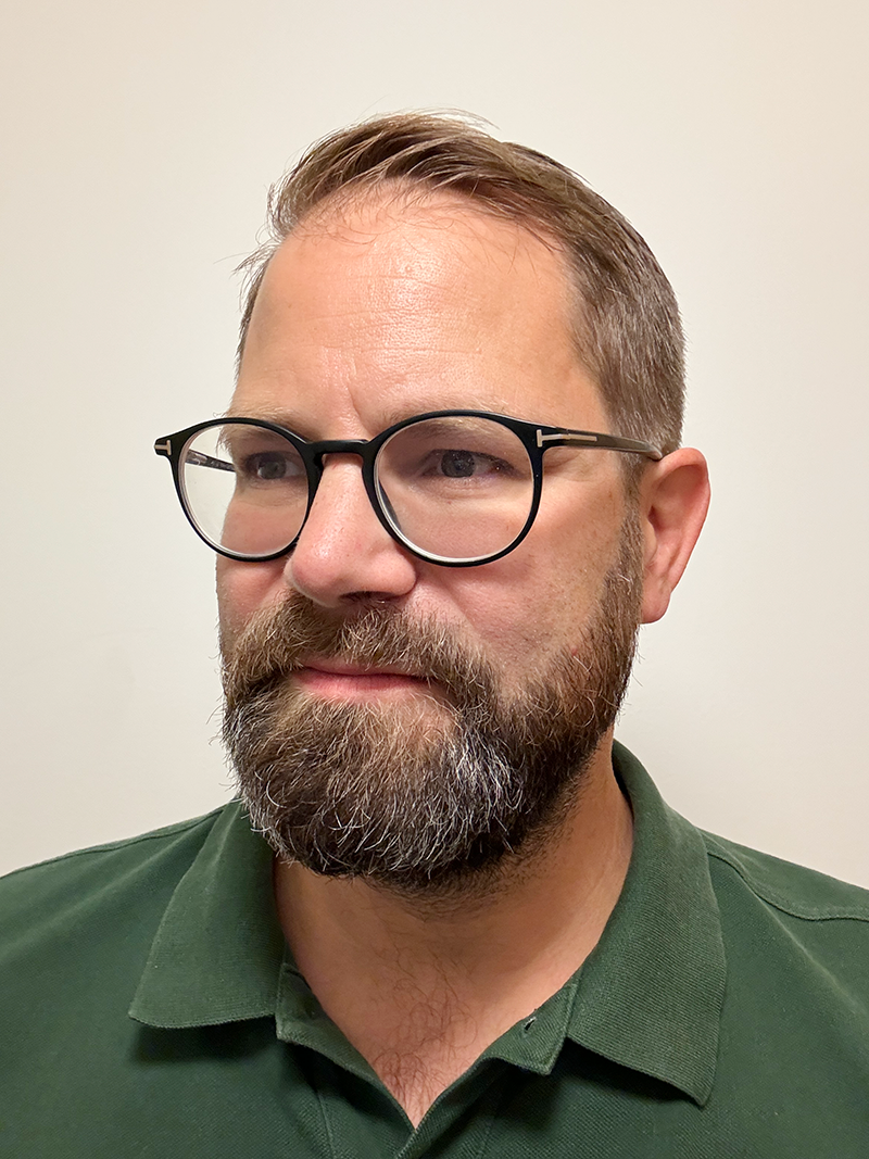 En man med kort brunt hår, skägg och glasögon har på sig en grön skjorta och står mot en ljus bakgrund.