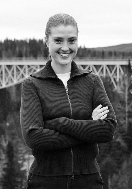 En kvinna står utomhus med armarna i kors och tittar mot kameran. En bro och träd syns i bakgrunden.