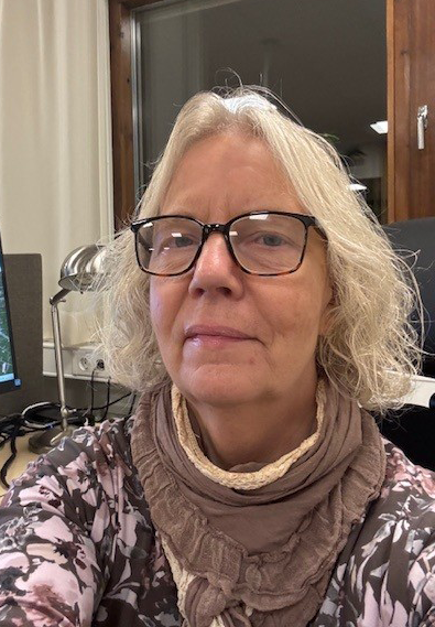En äldre kvinna med glasögon och axellångt grått hår sitter inomhus, har ett mönstrat plagg och en brun sjal, med skrivbord och dator i bakgrunden.
