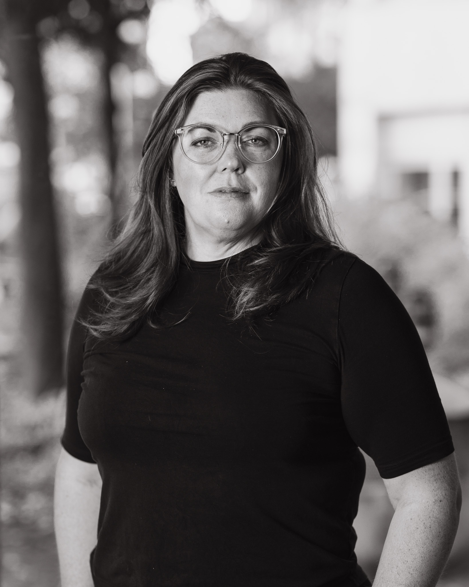 En kvinna med långt hår och glasögon har på sig en svart tröja och står utomhus, tittar mot kameran med neutral min. Bakgrunden är suddig.