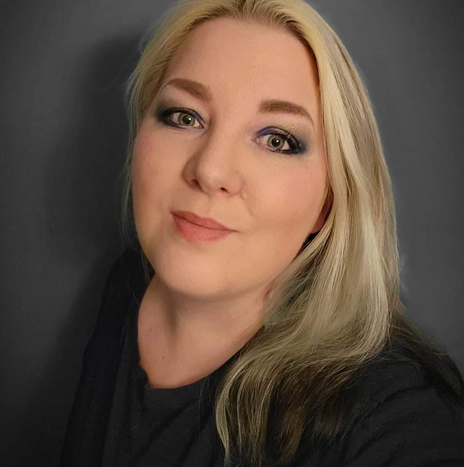 En kvinna med långt blont hår och mörka rötter, har mörk ögonmakeup och svart topp, poserar framför en mörk bakgrund.