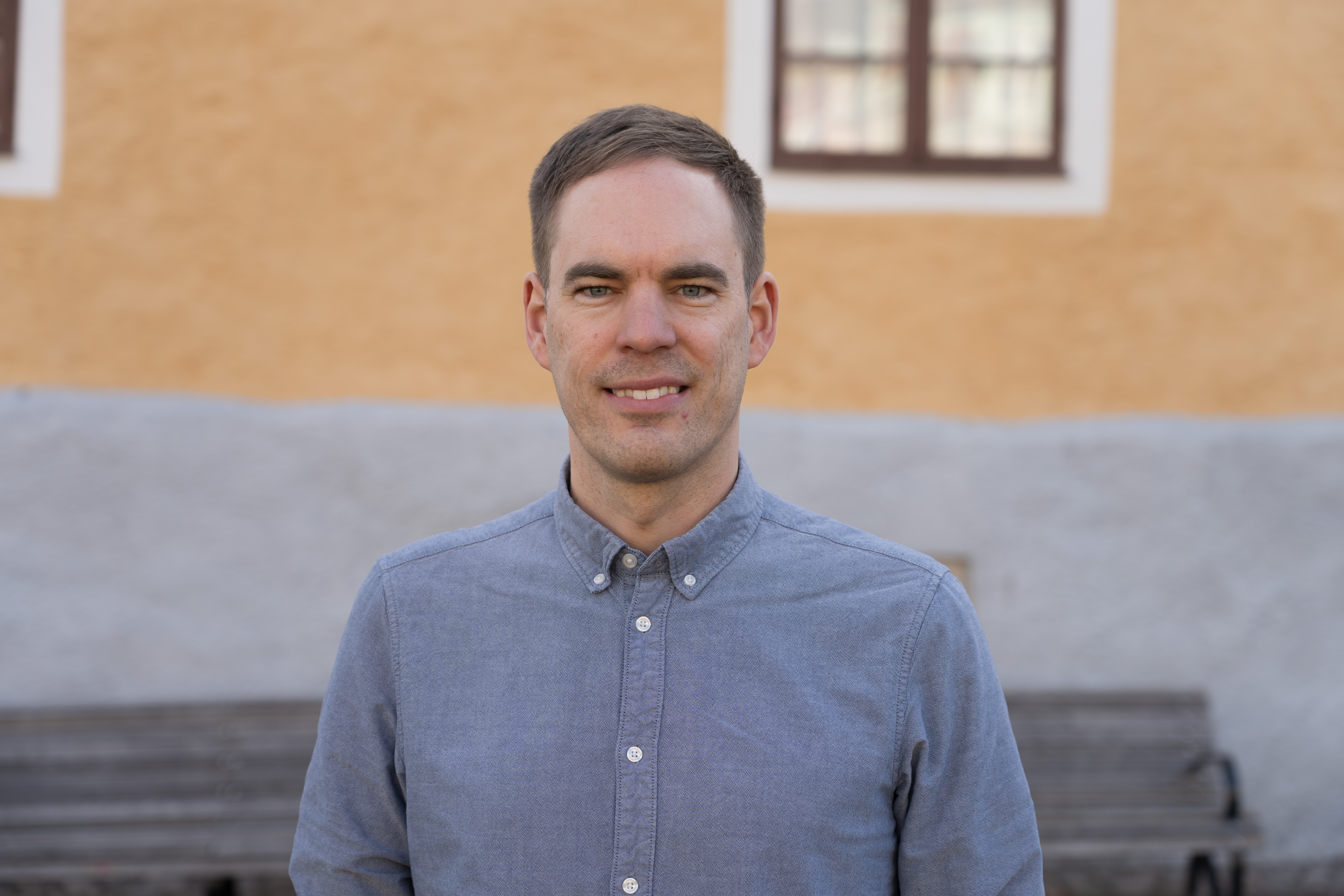 En man med kort brunt hår och ljusblå skjorta står utomhus framför en gul och grå byggnad.