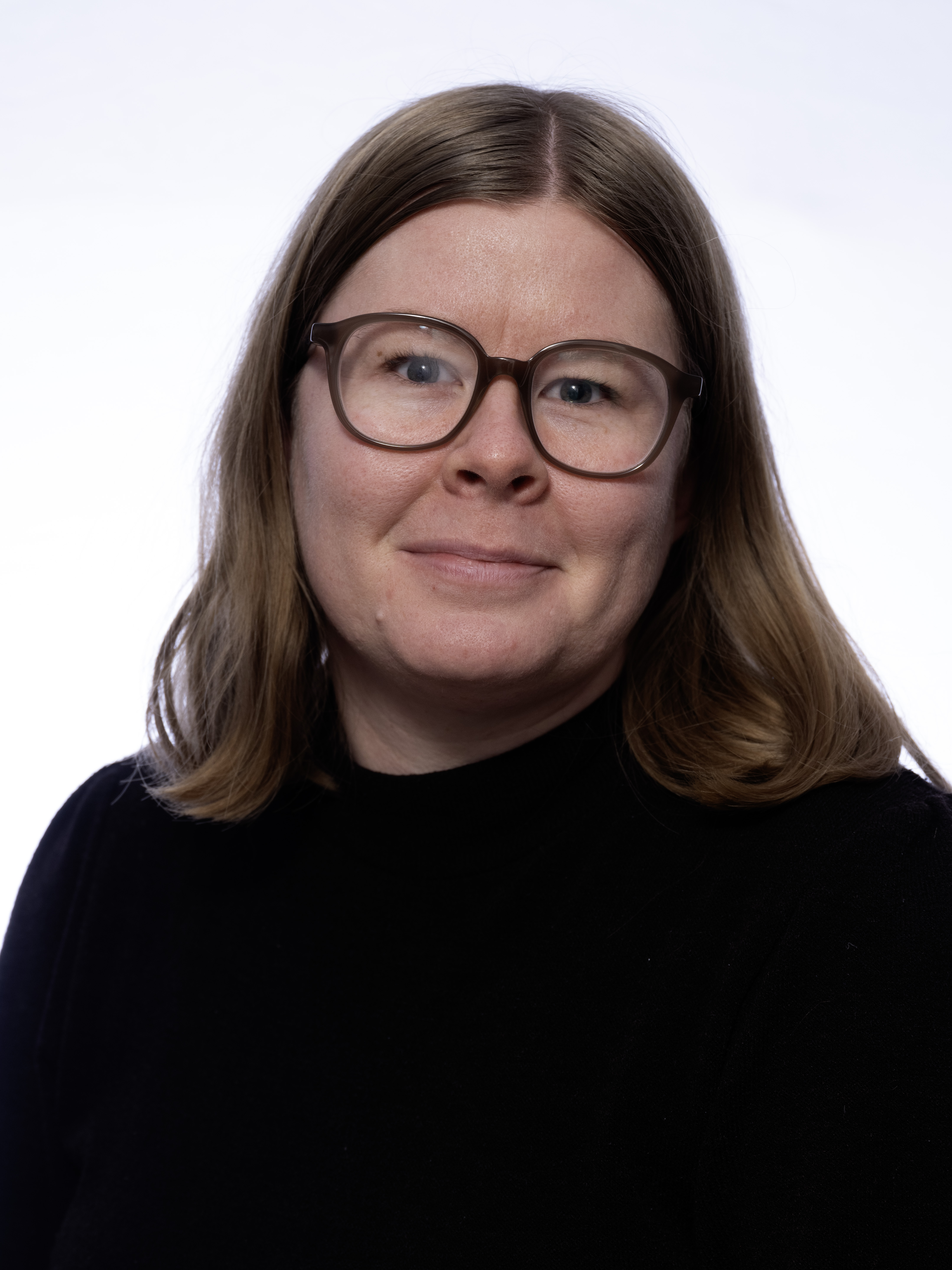 En person med axellångt brunt hår, glasögon och svart topp står framför en vit bakgrund.