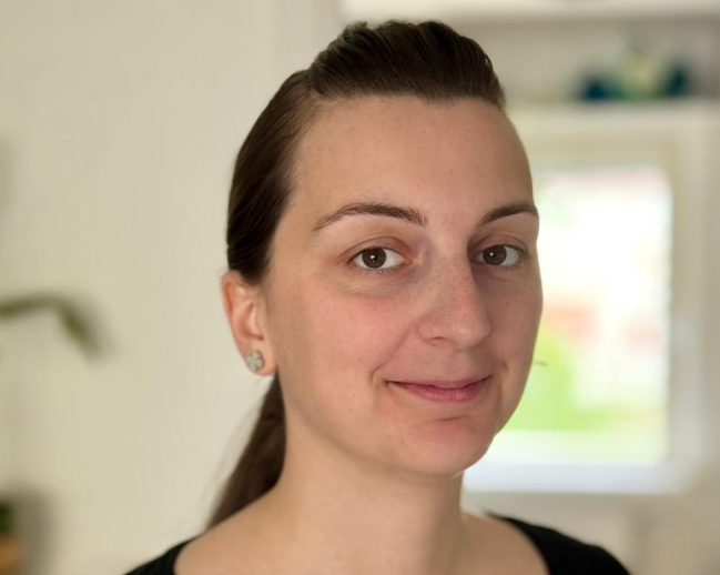 En kvinna med uppsatt brunt hår, svart topp och små örhängen står inomhus framför en suddig bakgrund.