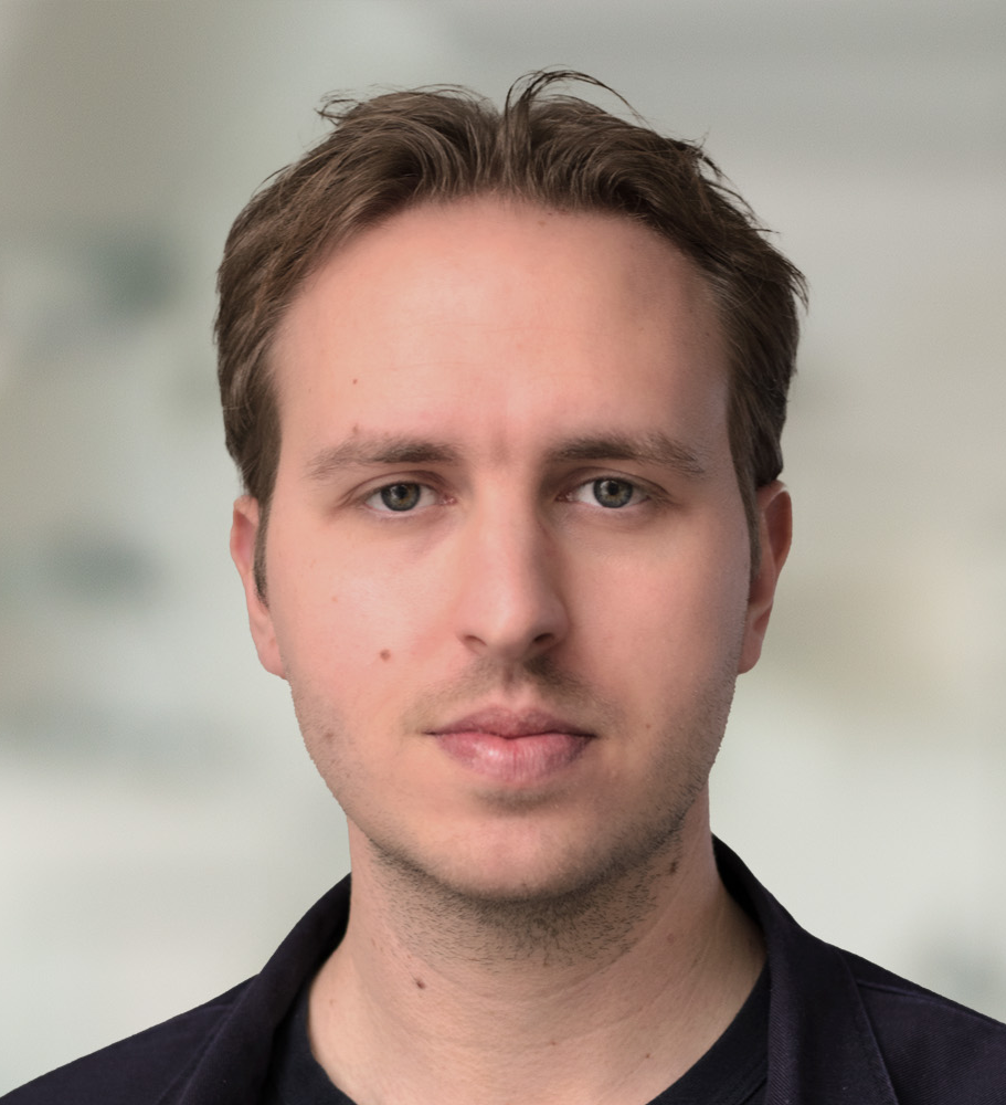 En man med kort brunt hår och ljus hud ser mot kameran med neutral min. Bakgrunden är suddig och visar en inomhusmiljö.