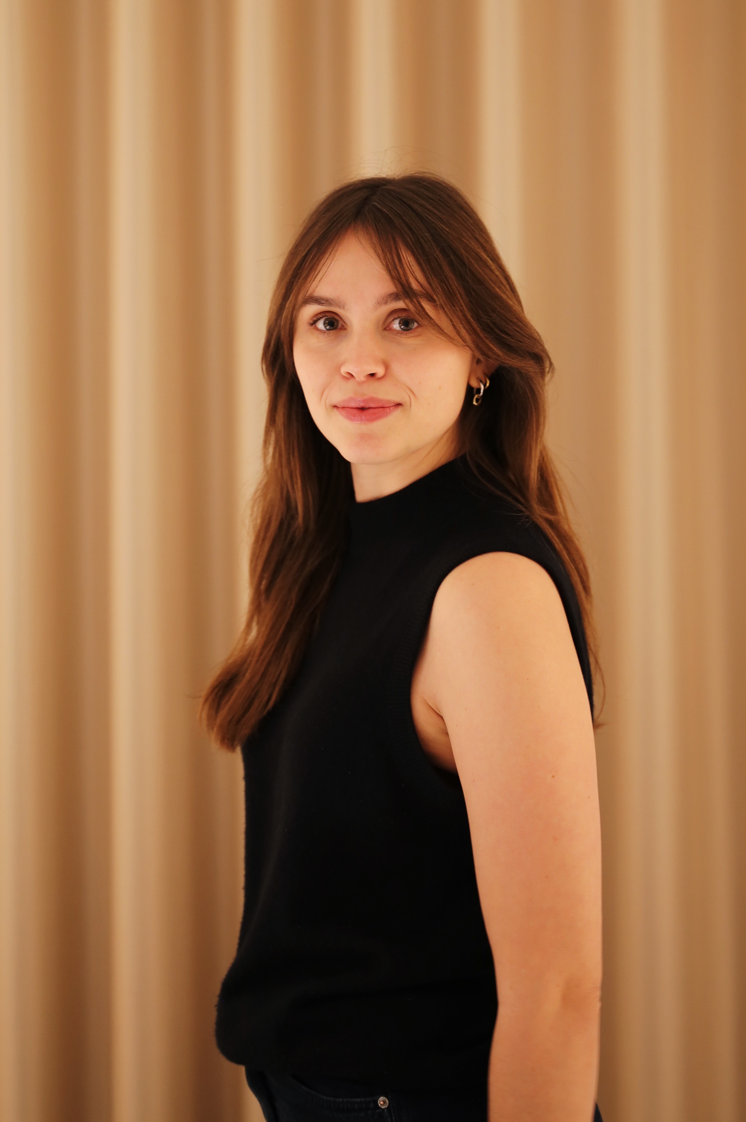 En kvinna med långt brunt hår och ärmlös svart topp står framför beige gardiner och tittar mot kameran med neutral min.