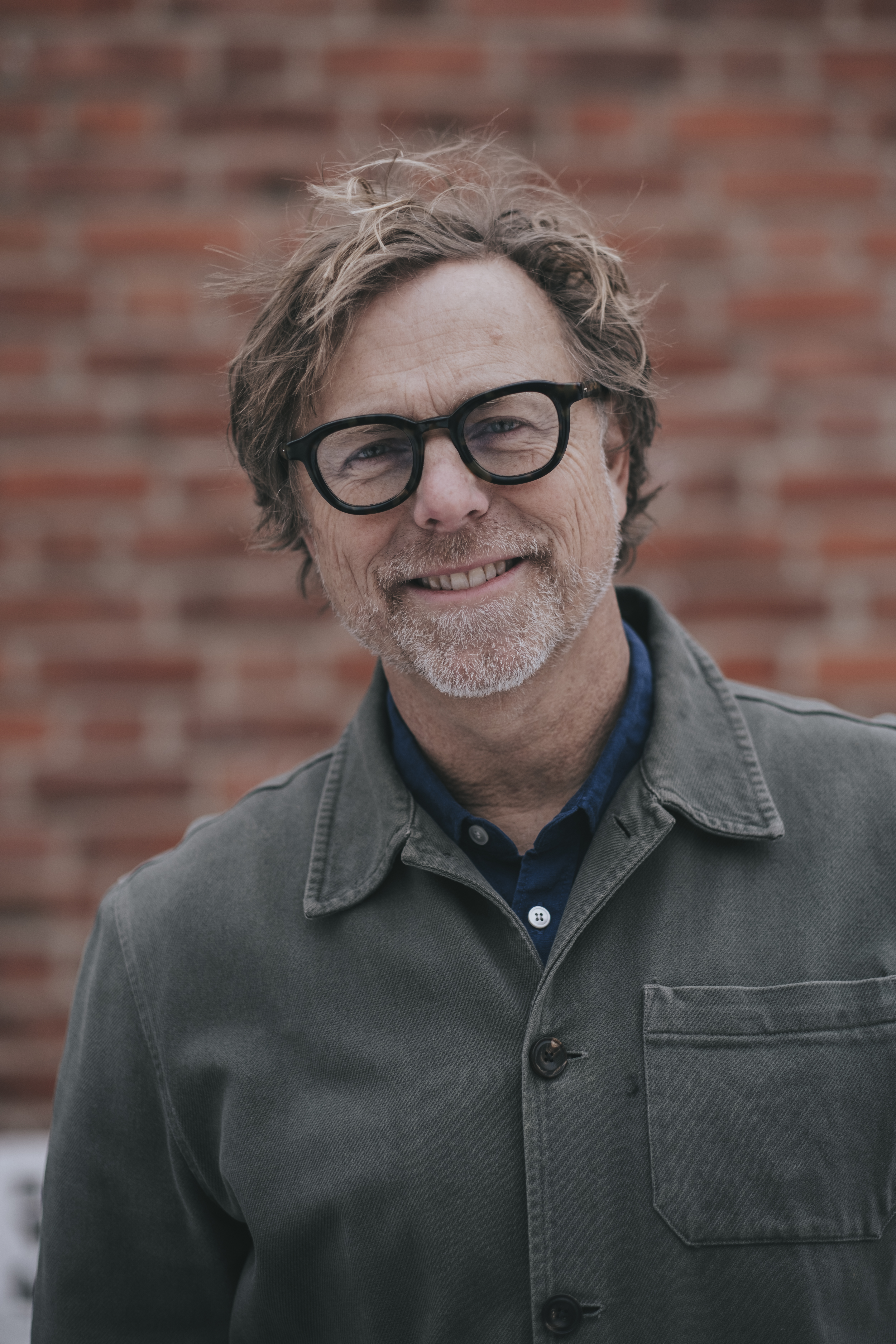 En medelålders man med glasögon, lätt skägg och rufsigt hår ler, bär en grå jacka och blå skjorta, står framför en tegelvägg.