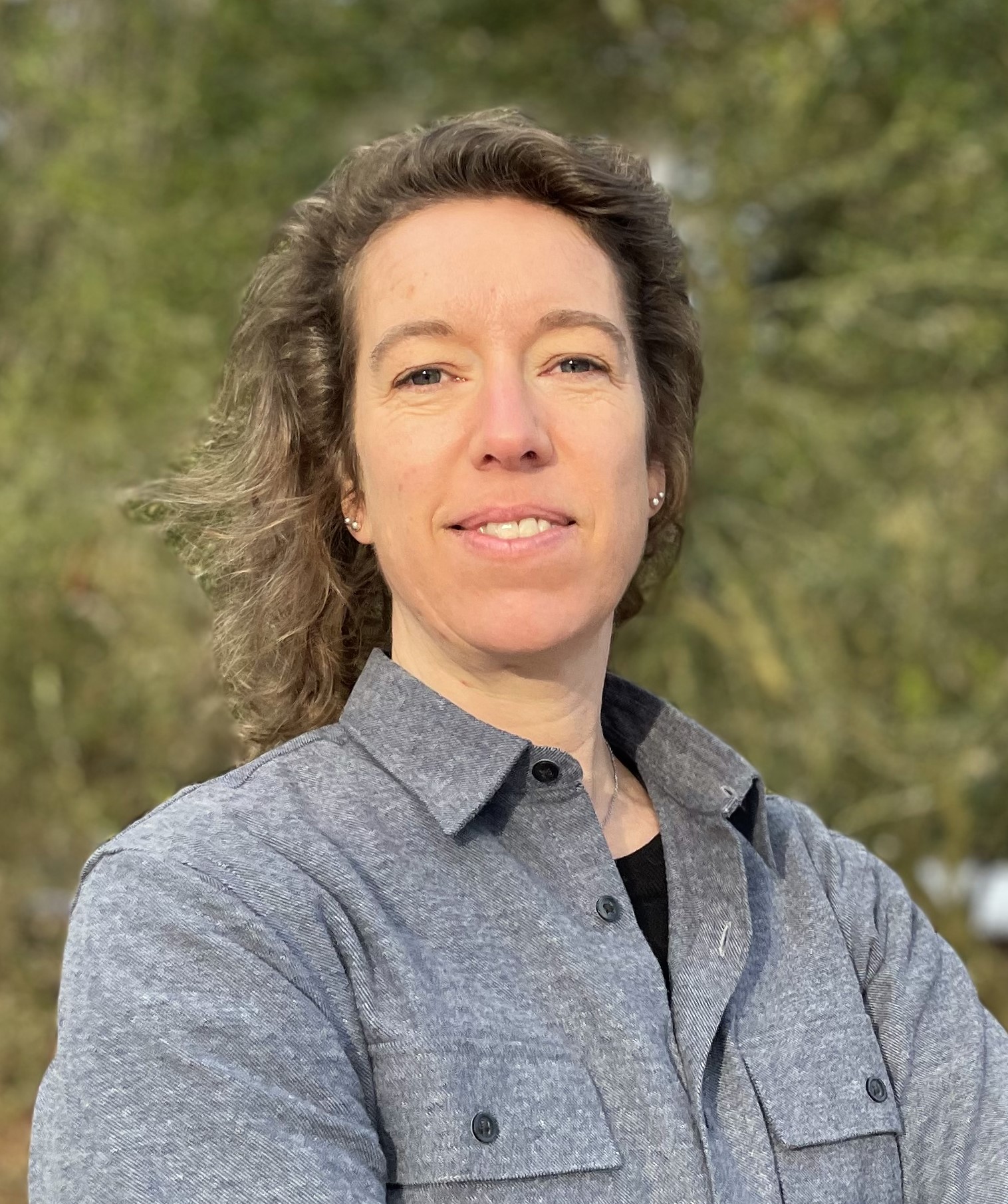 En person med vågigt brunt hår står utomhus med armarna i kors och har på sig en grå skjorta. Träd och växter syns bakom.