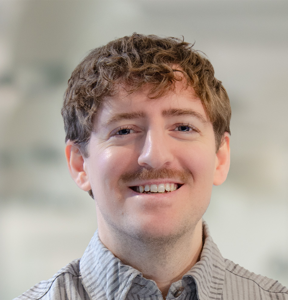 En man med ljust brunt lockigt hår och mustasch ler mot kameran. Han har på sig en ljus skjorta. Bakgrunden är suddig.