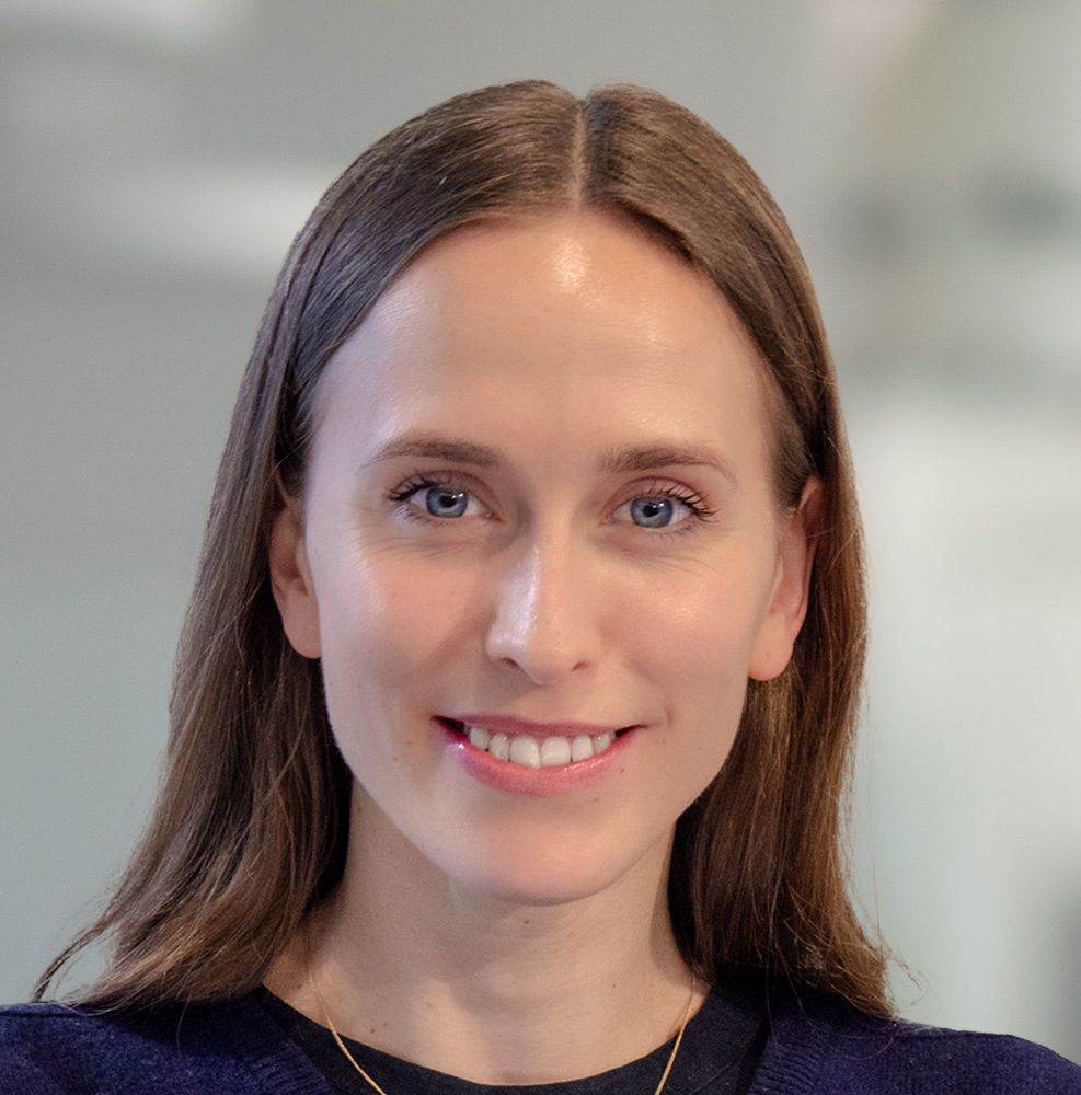 En kvinna med rakt brunt hår och blå ögon ler, har på sig en mörk topp och ett guldhalsband i en inomhusmiljö.