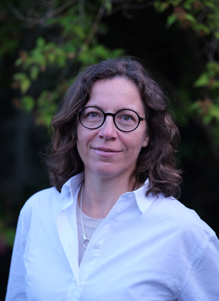 En kvinna med brunt axellångt hår och glasögon, iklädd en vit skjorta, står utomhus med gröna växter i bakgrunden.