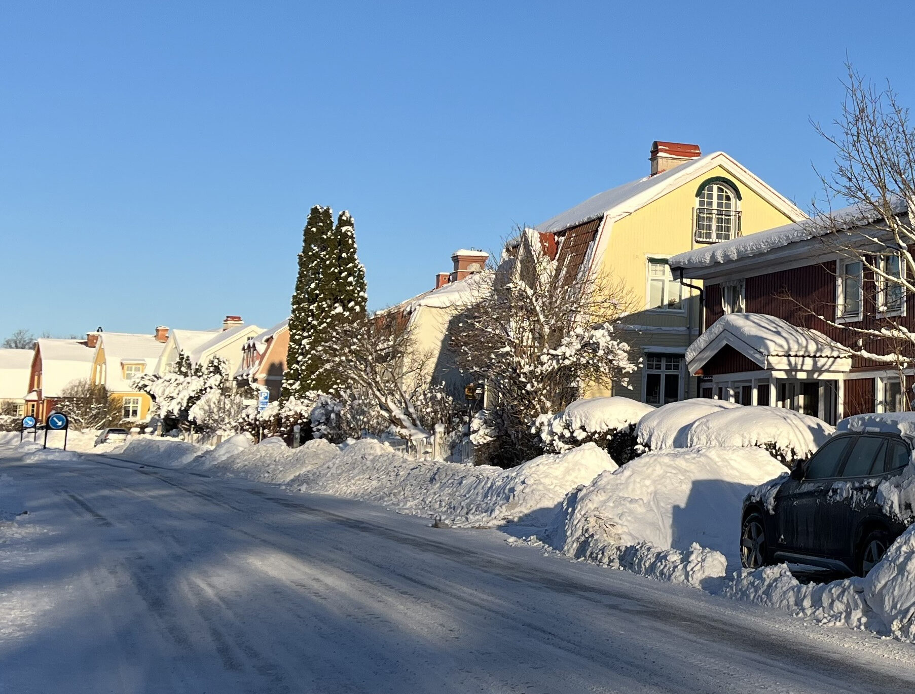 Bostadsgata med snö, hus, träd och parkerade bilar under blå himmel en vinterdag.