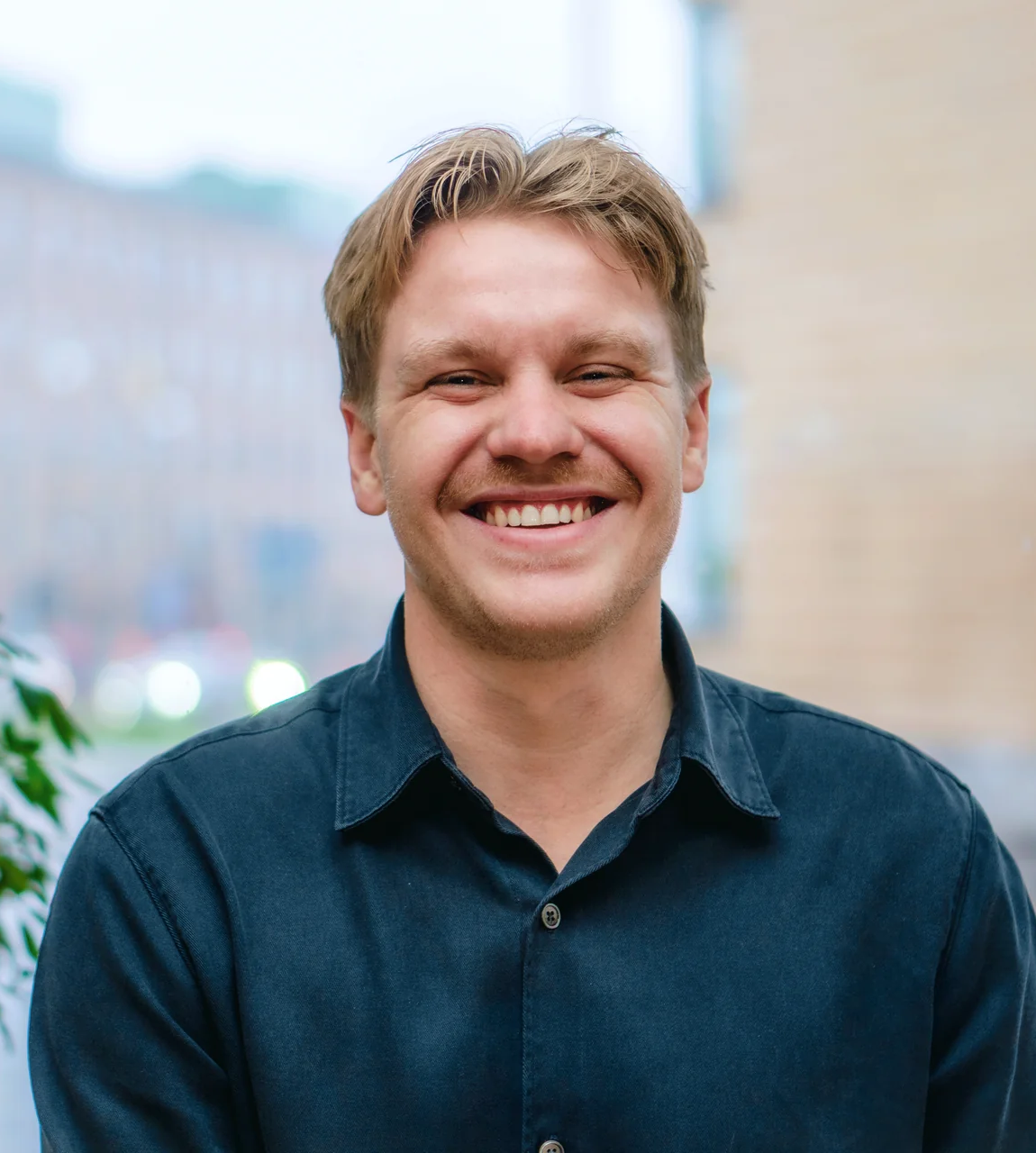 En man med ljusbrunt hår och mustasch, i mörk skjorta, ler inomhus med en suddig stadsmiljö i bakgrunden.