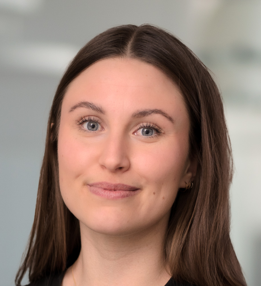 En kvinna med långt brunt hår och ljusa ögon, klädd i svart topp, tittar in i kameran mot en suddig bakgrund inomhus.