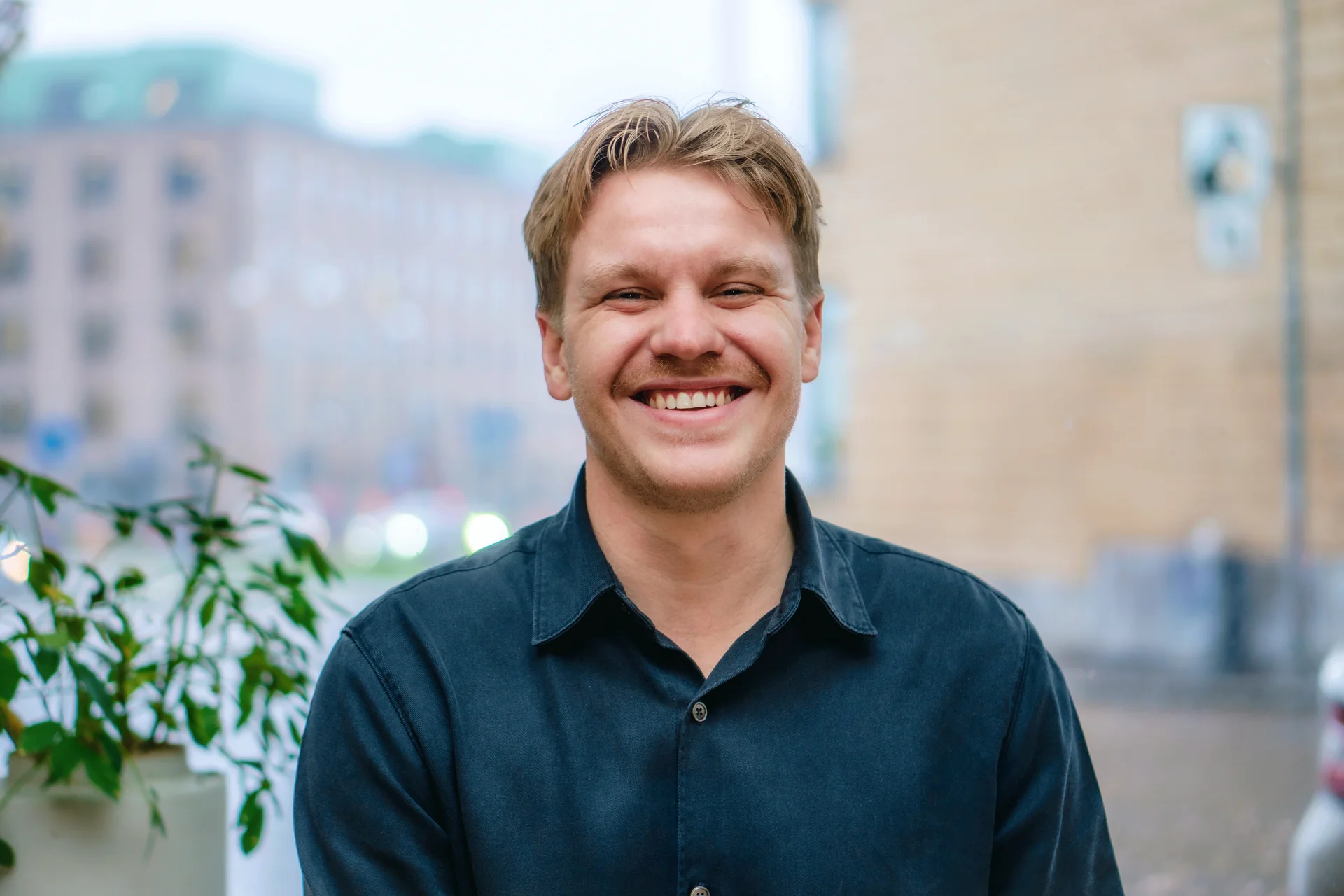 En man med ljust brunt hår och mustasch ler mot kameran, har en mörk skjorta på sig, stadens byggnader syns i bakgrunden.