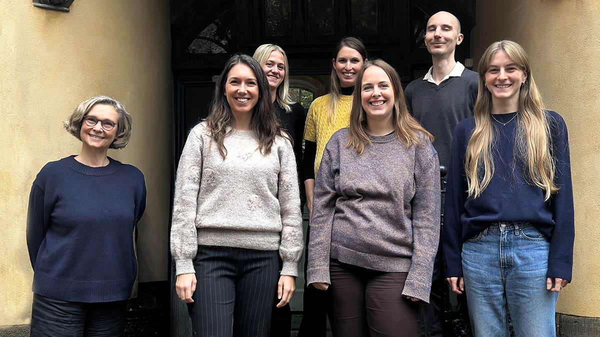 Sju personer står framför en byggnadsentré och poserar för ett gruppfoto, alla har avslappnade kläder och ler.