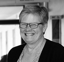 En medelålders person med kort hår och glasögon som ler, har på sig en mönstrad topp och mörk kofta, inomhus.