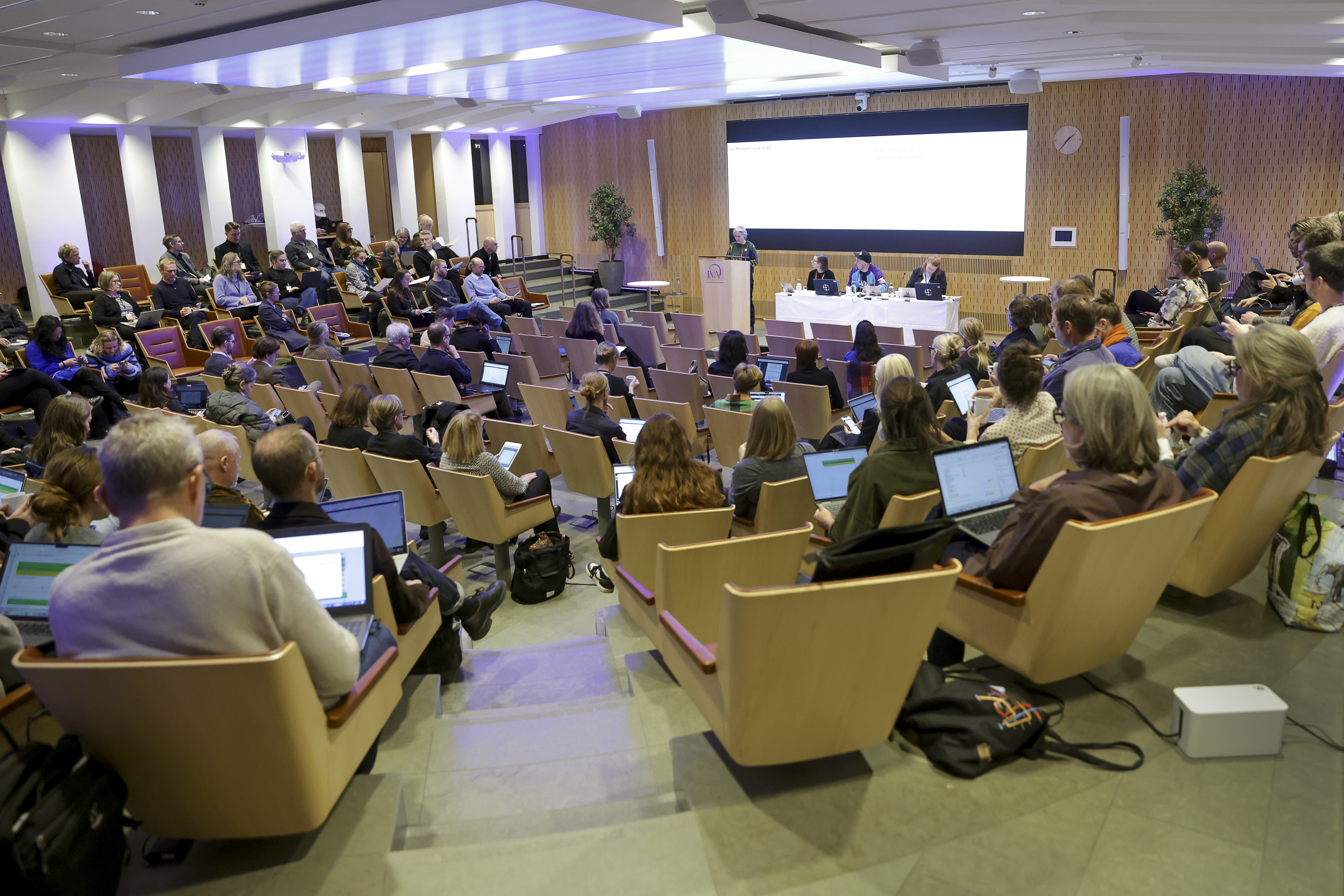 Personer sitter i en aula och lyssnar på en paneldiskussion längst fram, många använder datorer och antecknar.