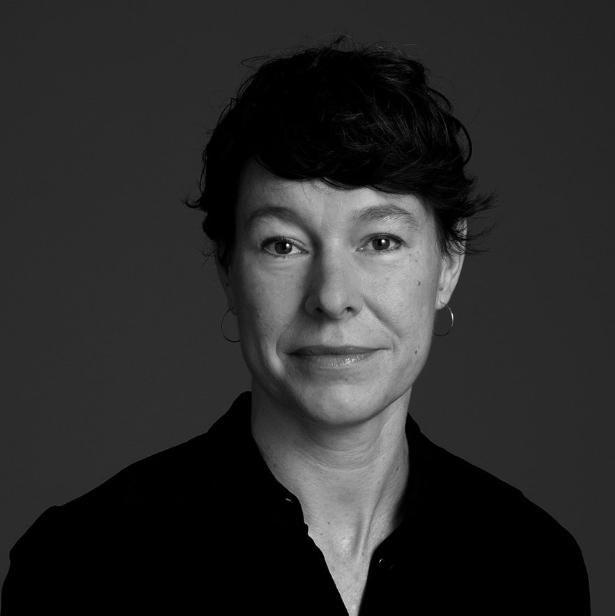 Porträtt i svartvitt av en kvinna med kort mörkt hår, hon har hoops i öronen och en mörk skjorta, tittar mot kameran.