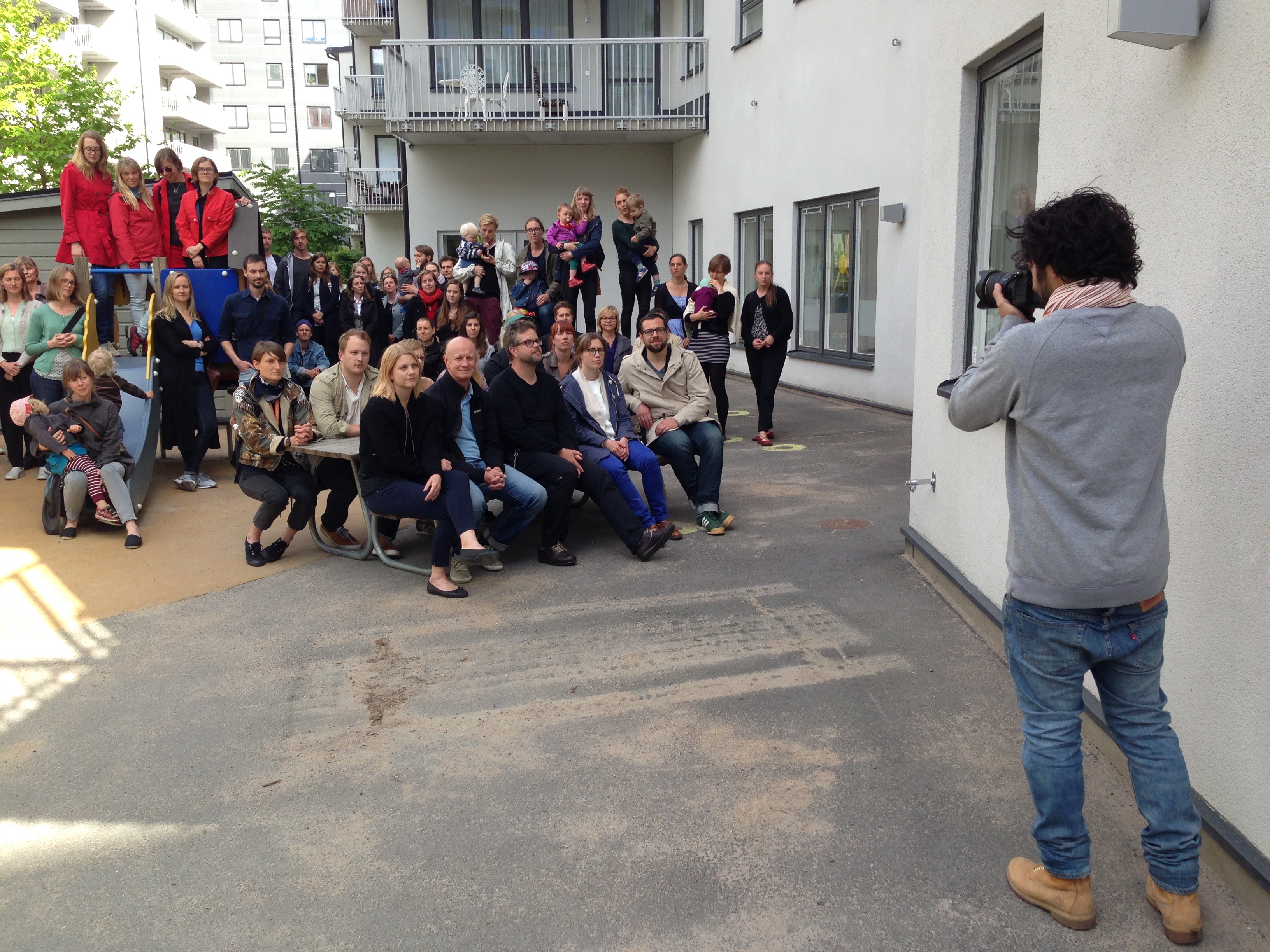 En fotograf tar ett gruppfoto av personer i olika åldrar som sitter och står utanför ett bostadshus.
