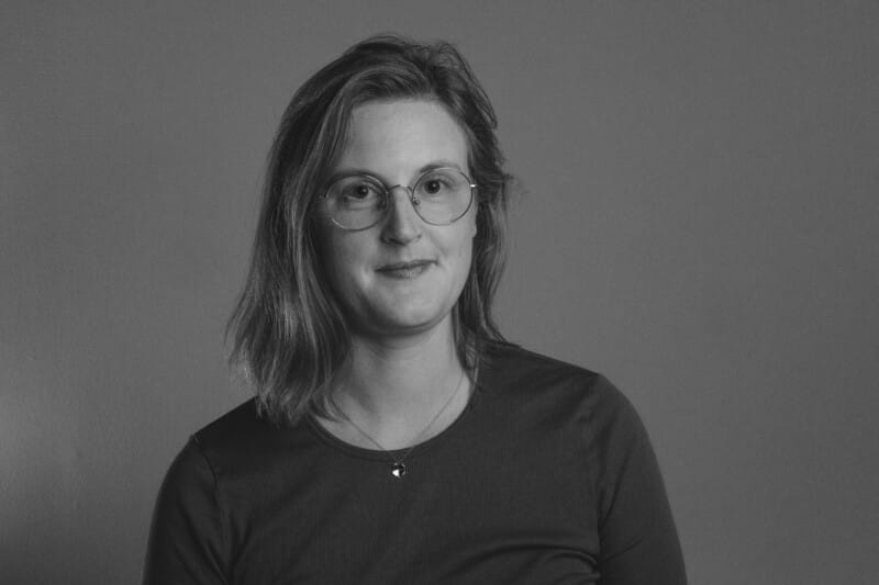 En kvinna med axellångt hår och glasögon har en enkel tröja på sig och tittar in i kameran mot en enfärgad bakgrund. Svartvitt foto.
