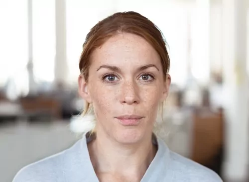 En kvinna med ljus hud och rödbrunt hår tittar rakt mot kameran i ett kontorsmiljö.