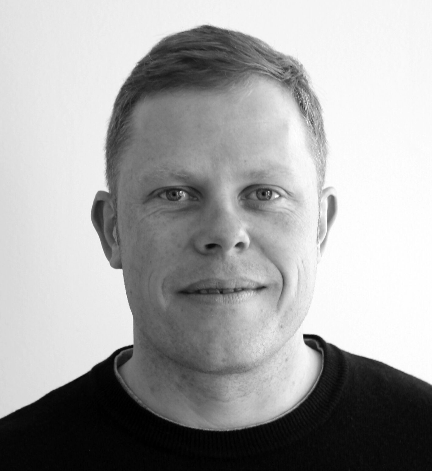 En man med kort ljust hår och ett svagt leende ser mot kameran framför en vit bakgrund. Han har en mörk tröja på sig.
