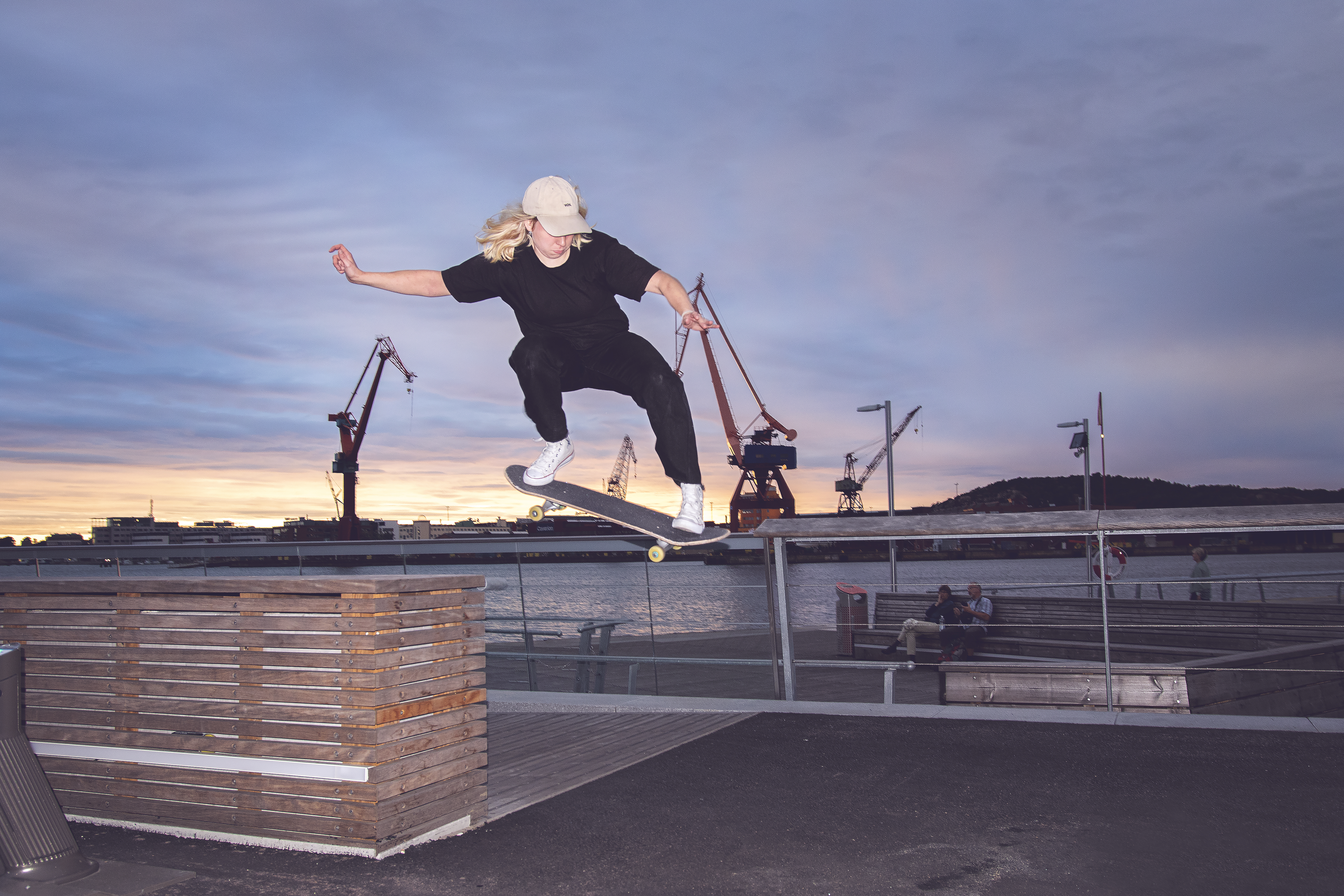 En person med keps och svarta kläder gör ett skateboardtrick vid en kaj. I bakgrunden syns kranar och vatten.