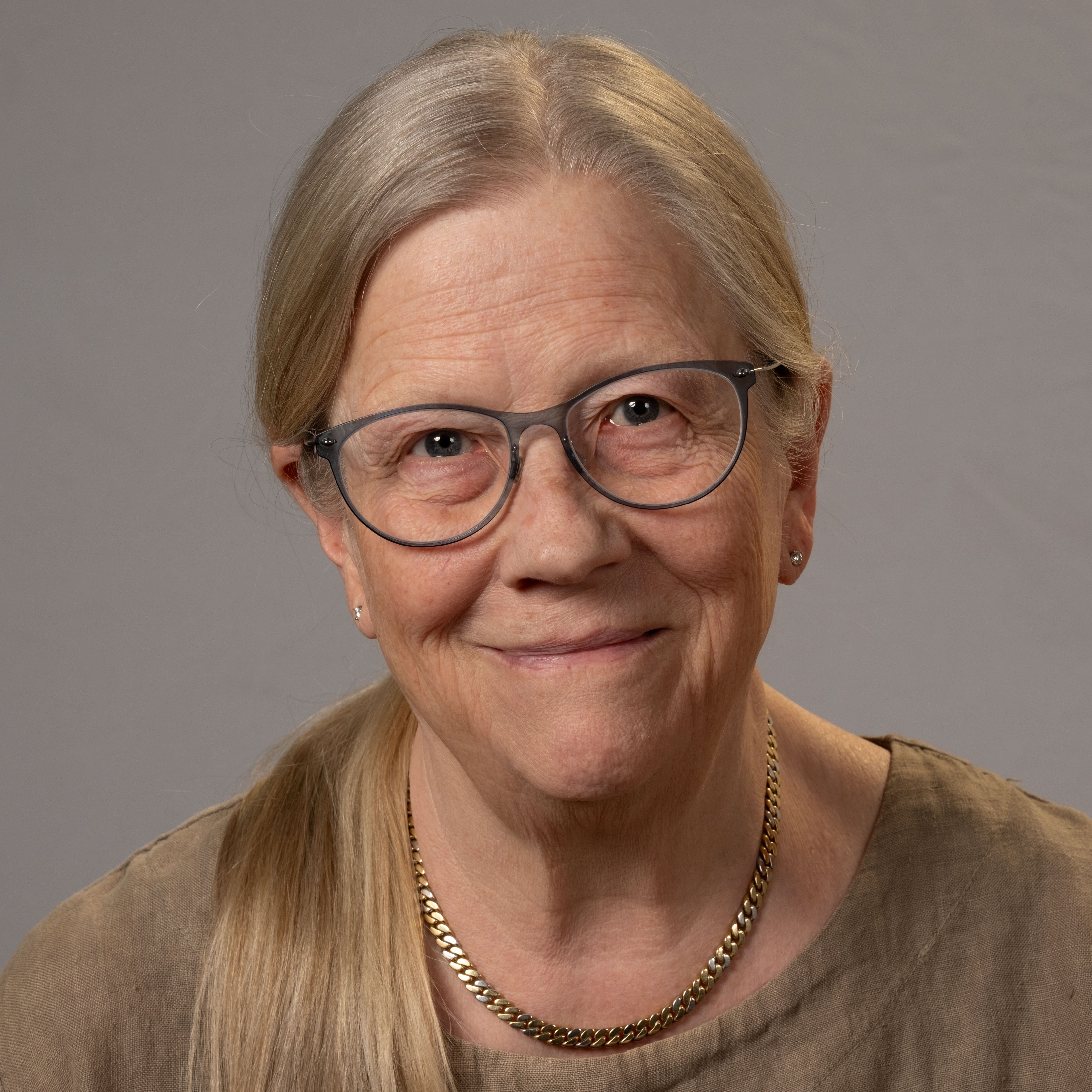 En äldre kvinna med långt grått hår, glasögon och ett guldfärgat halsband ler mot kameran. Hon har en ljusbrun topp. Bakgrunden är grå.
