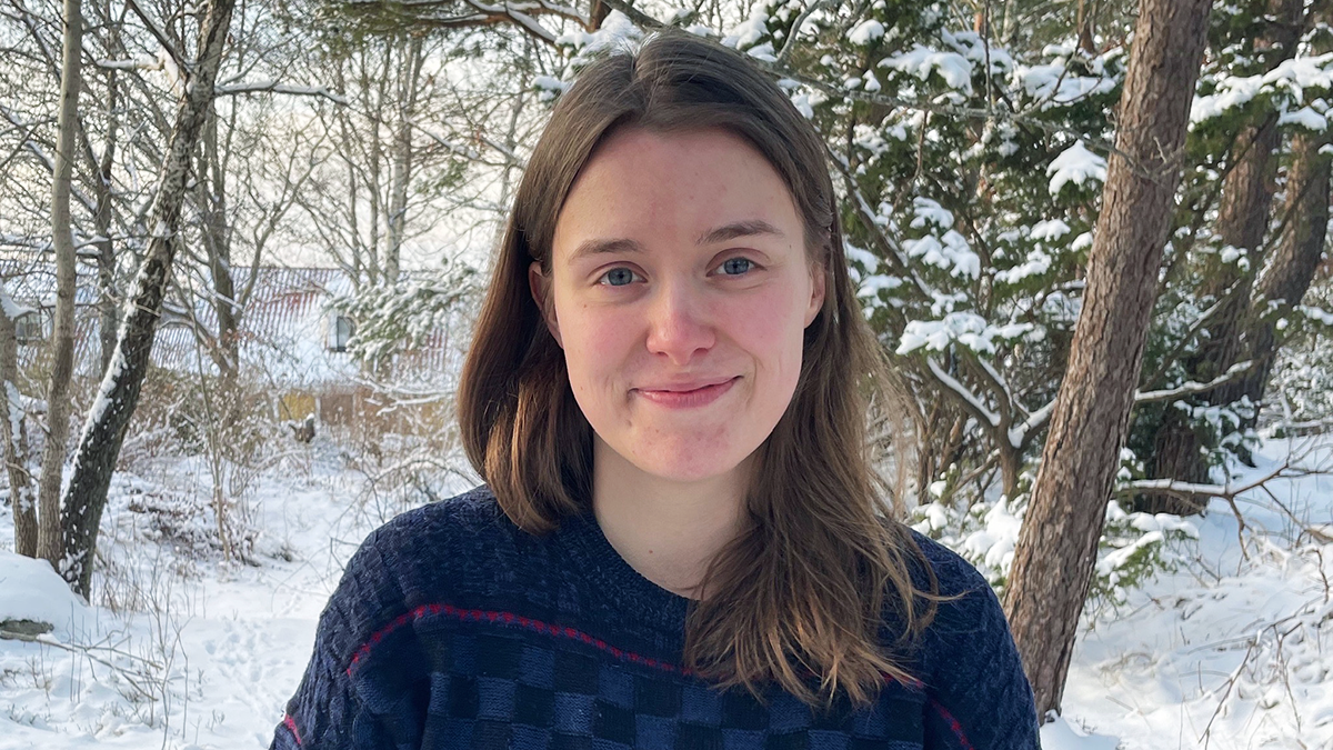 En person med långt brunt hår och en mörkrutig tröja står utomhus i ett snötäckt skogsområde och tittar mot kameran.