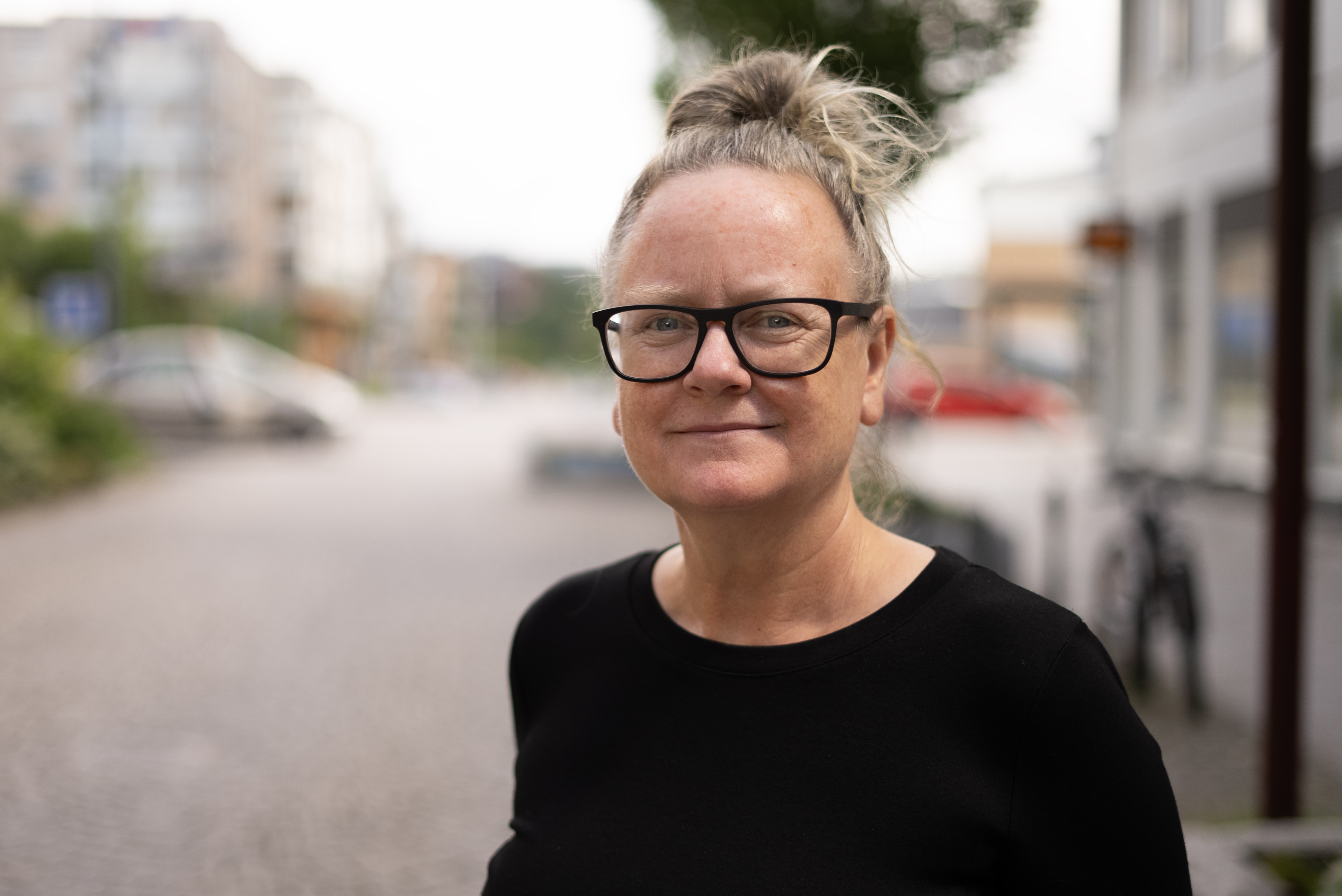 En person med glasögon och håret i en knut står utomhus på en kullerstensgata med svarta tröja. Hus och bilar syns bakom.