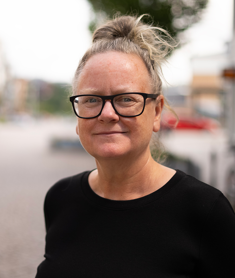 En person med ljus hud, glasögon och grått hår i en knut har en svart tröja och står utomhus på en stadsgata.