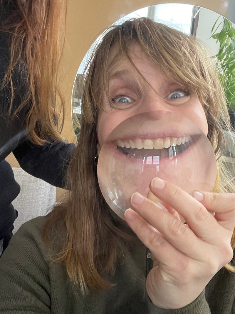 En kvinna håller ett stort, runt förstoringsglas framför munnen så att leendet ser förändrat ut. En annan person står nära.