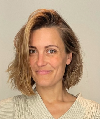 En kvinna med kort, vågigt, ljusbrunt hår och ljus hud bär en stickad tröja och ler svagt framför en enkel bakgrund.