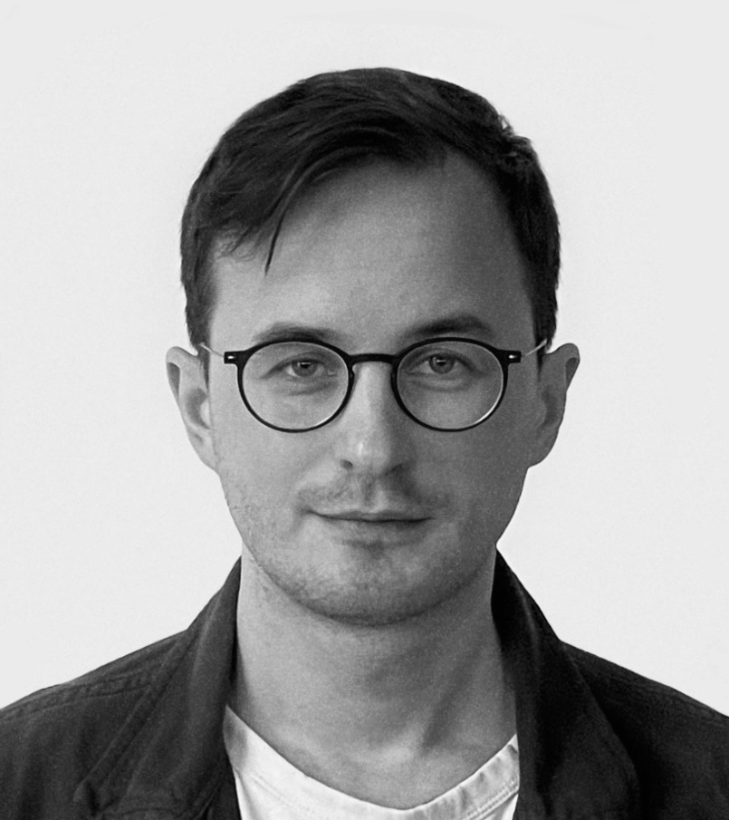 En person med kort mörkt hår, runda glasögon, vit skjorta och mörk jacka ser in i kameran framför en enkel bakgrund.