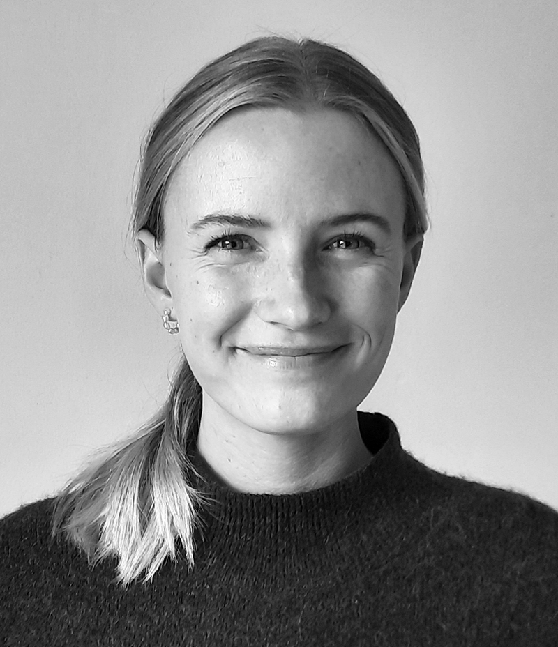 En kvinna med ljust hår i en sidotoffs, har på sig en mörk tröja och ler mot kameran framför en enkel bakgrund.