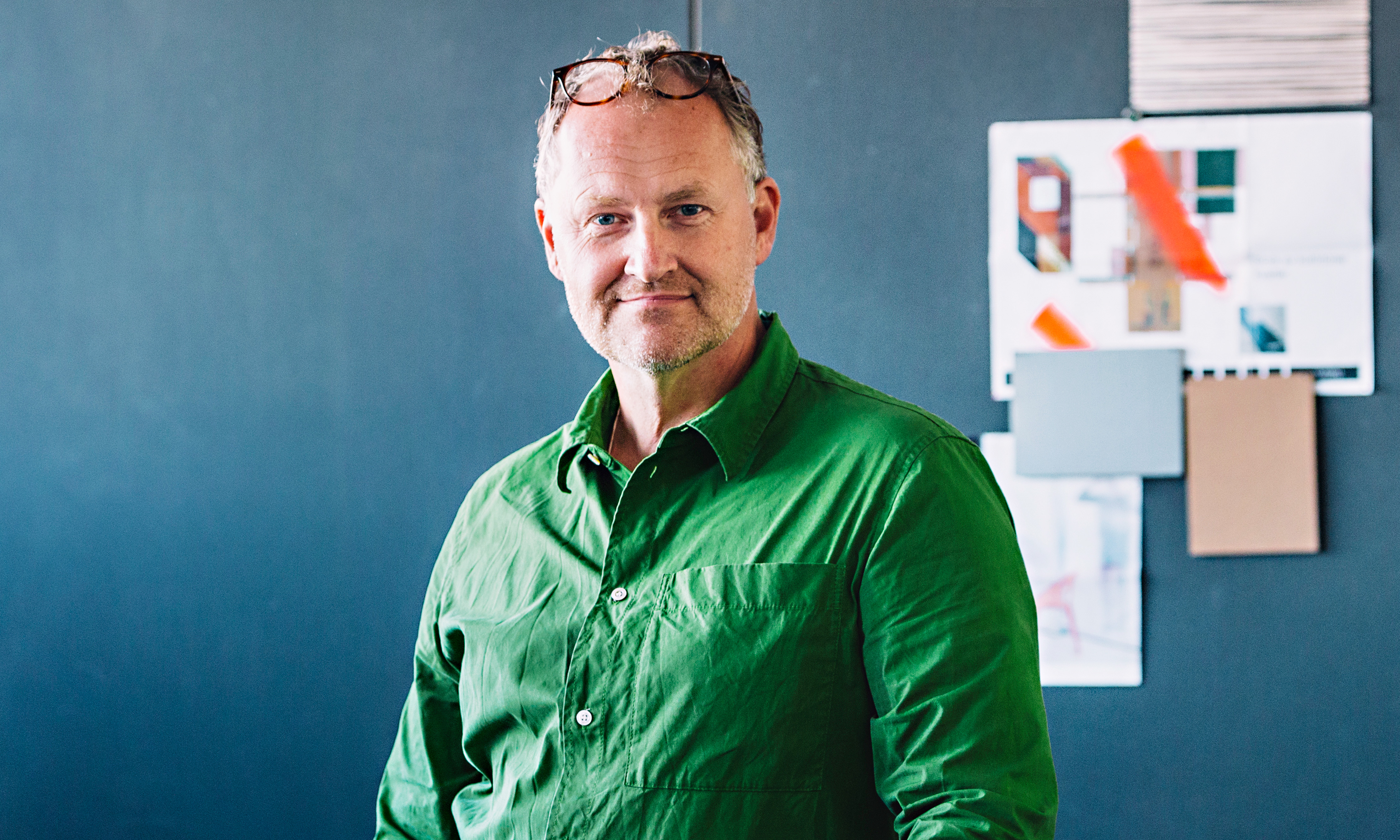 En man med grått hår och glasögon på huvudet, iklädd en grön skjorta, står framför en mörk vägg med designmaterial och färgprover bakom sig.