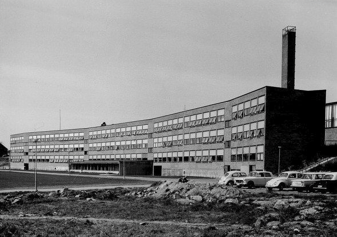 Svartvit bild av en flervåningsbyggnad med många fönster, flera äldre bilar står framför och en skorsten syns i ena änden.