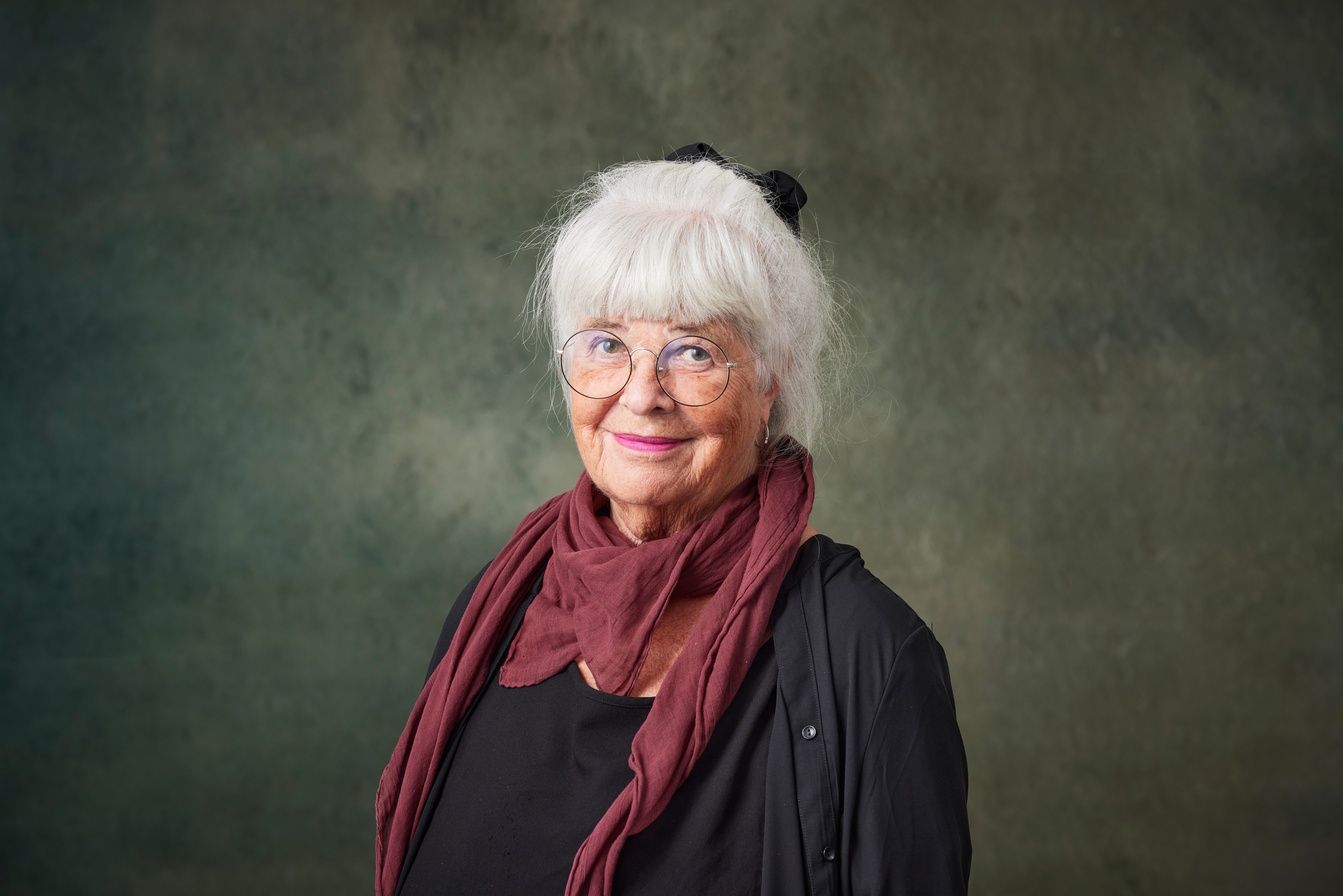 Äldre kvinna med vitt hår, glasögon och mörka kläder samt en vinröd scarf, står framför en enkel bakgrund.