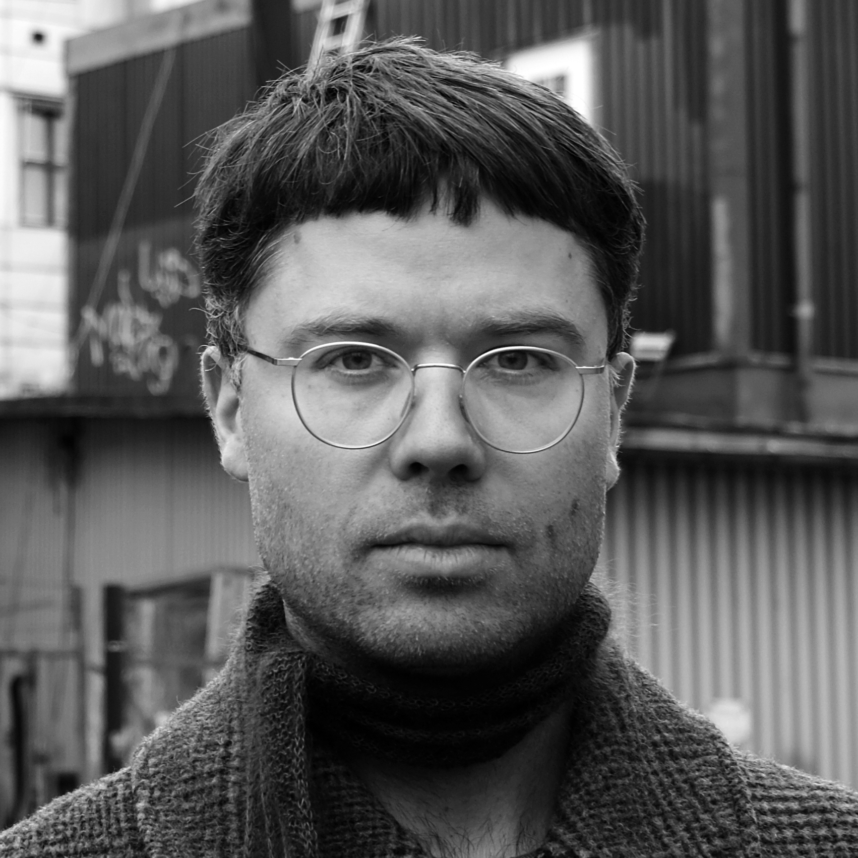 En man med kort mörkt hår och runda glasögon ser mot kameran, har på sig tröja och halsduk, i en stadsmiljö.