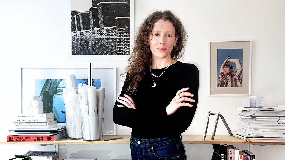 En kvinna med lockigt hår står med armarna i kors framför hyllor med böcker, papper och konsttryck i ett upplyst rum.