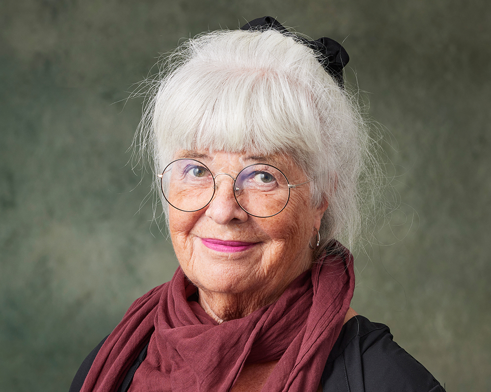 Äldre kvinna med vitt hår, runda glasögon och en mörkröd halsduk står framför en grön bakgrund.