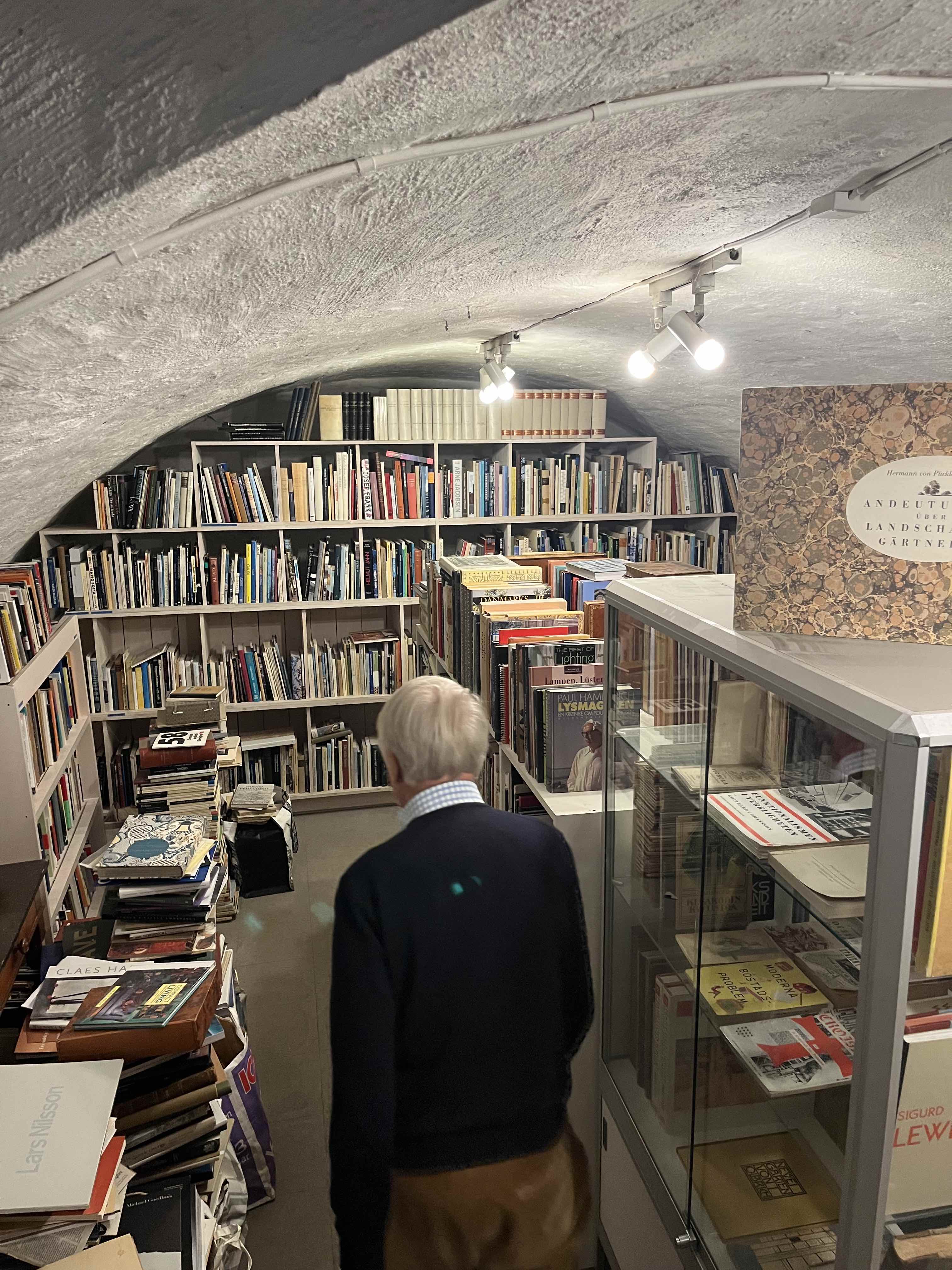 En äldre person står i ett bibliotek med välvda tak där hyllor håller böcker och dokument.