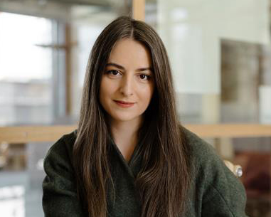 En kvinna med långt rakt brunt hår sitter inomhus, har på sig en mörkgrön tröja och tittar rakt mot kameran.