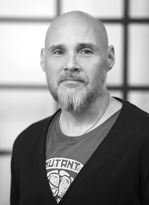 En skallig man med skägg och mustasch har på sig en t-shirt med tryck, kofta och halsband framför ett fönster. Svartvit bild.