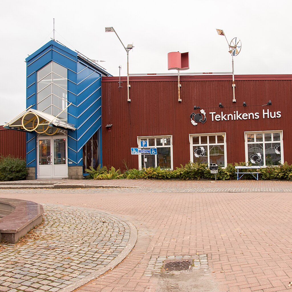 En röd byggnad med blå entré märkt "Teknikens Hus" har parkeringsskyltar, stora fönster och vindkraftverk på taket.