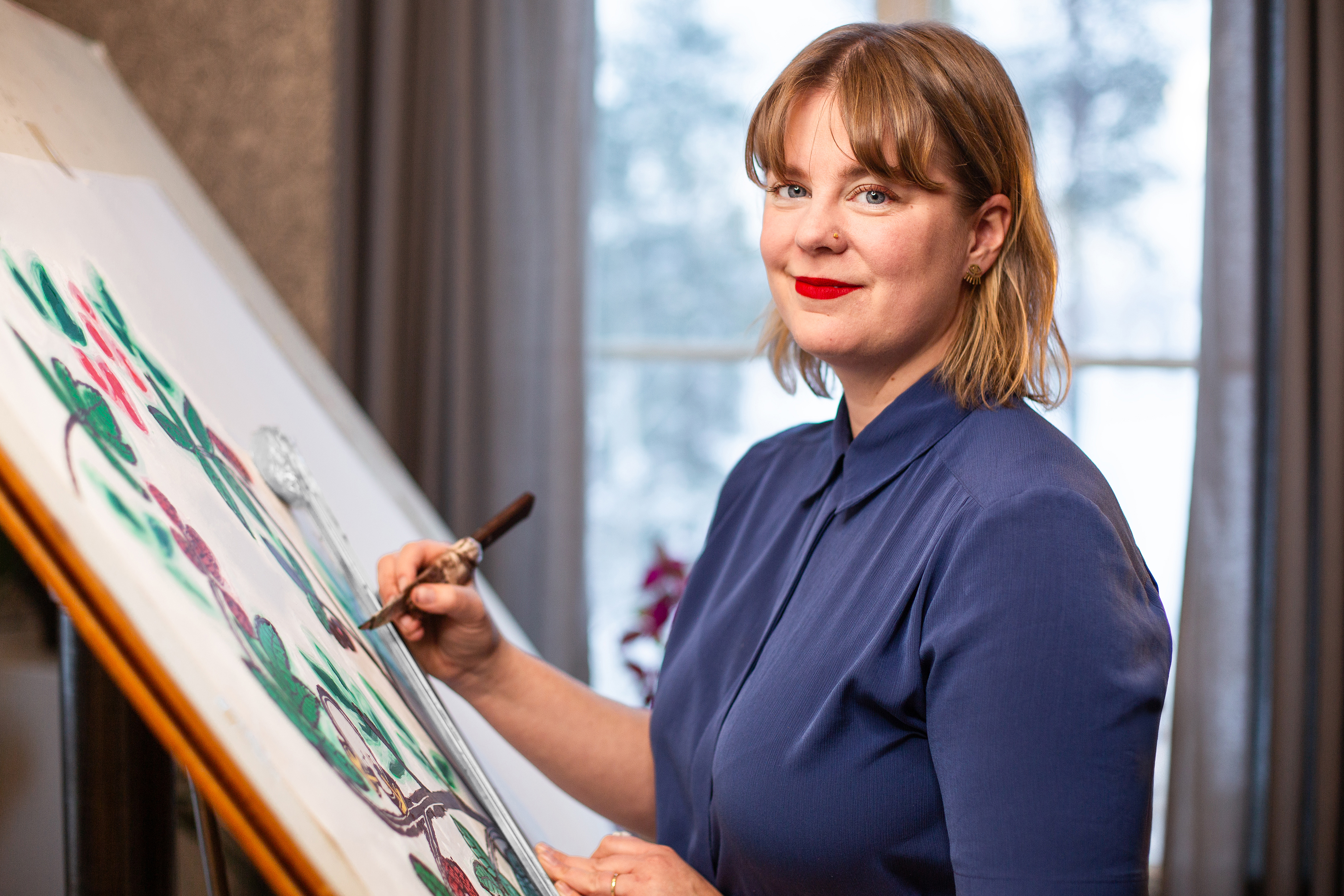 En kvinna i blå skjorta står vid ett staffli och målar med en pensel. Hon tittar mot kameran i ett rum med ett fönster bakom sig.
