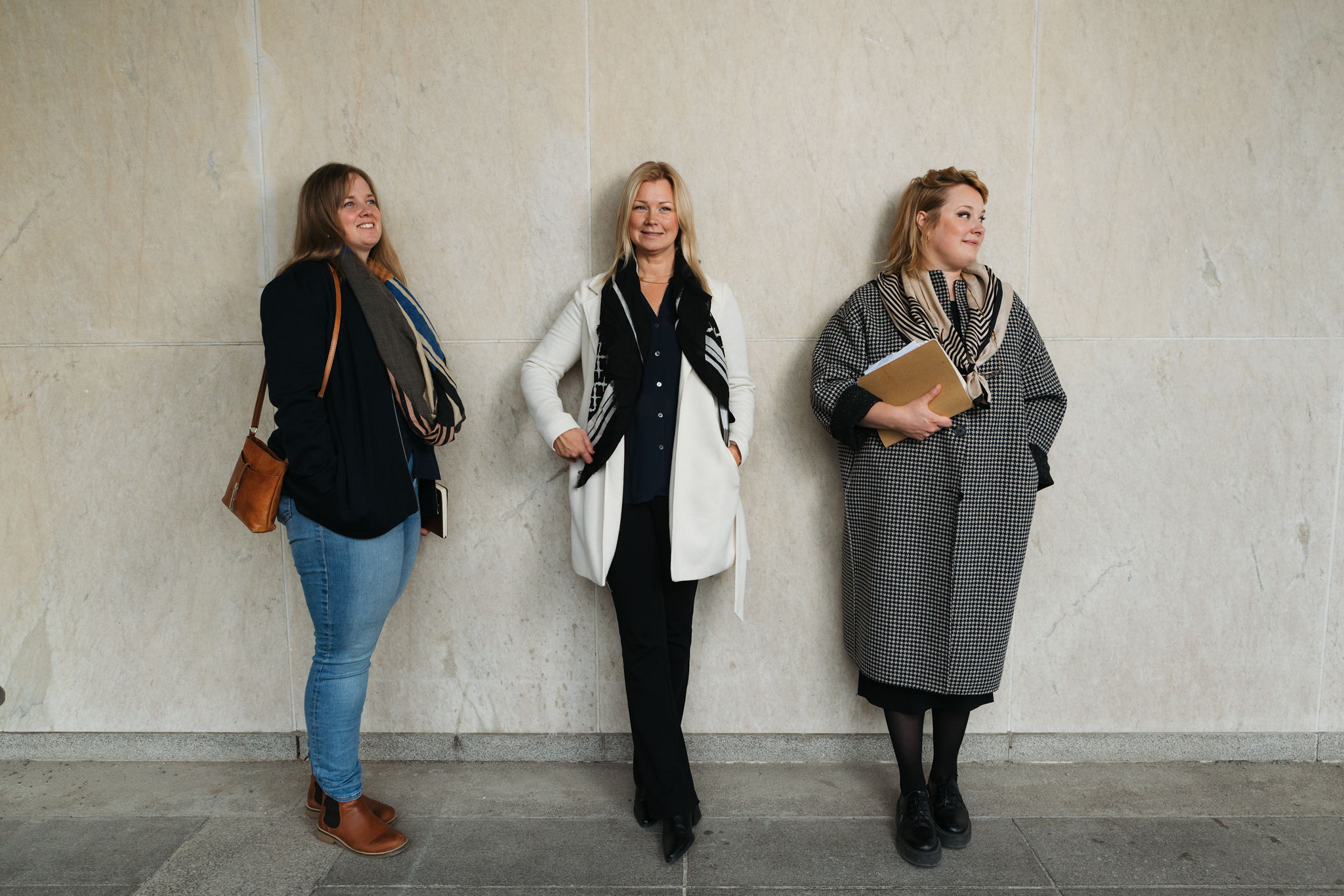 Tre kvinnor står mot en ljus vägg, klädda i business casual, en håller i en mapp, alla tittar åt olika håll.