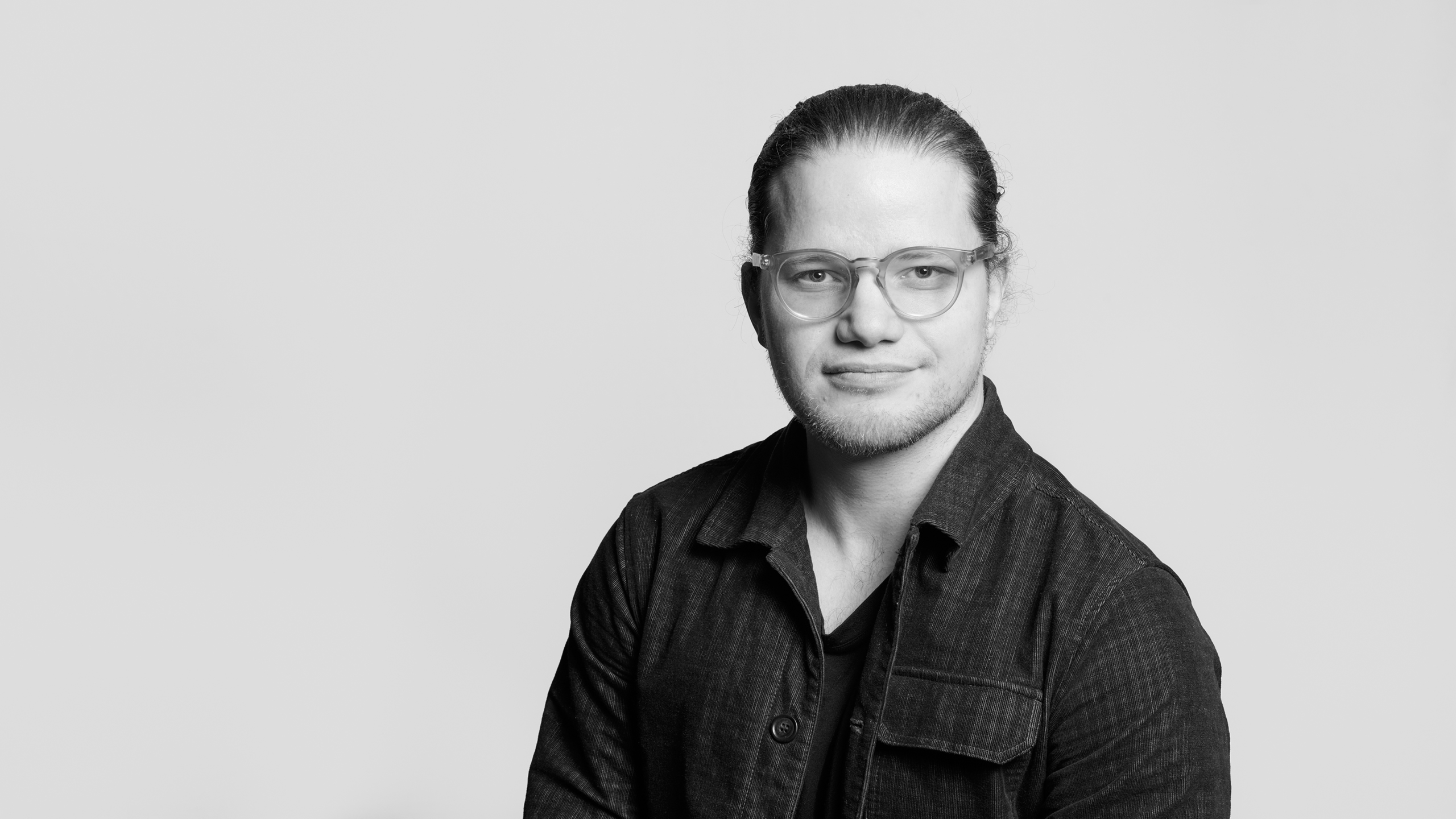 En person med glasögon och långt hår uppsatt, har en mörk jacka över en t-shirt och står mot en ljus bakgrund.