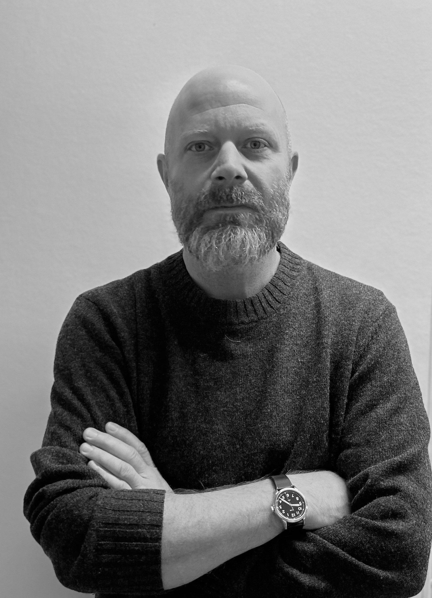 En man med skägg står mot en enkel bakgrund med armarna i kors, bär en mörk tröja och en handledsklocka.